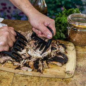 Black meat claws shred pulled pork on a wooden cutting board. A spice jar and parsley are nearby.