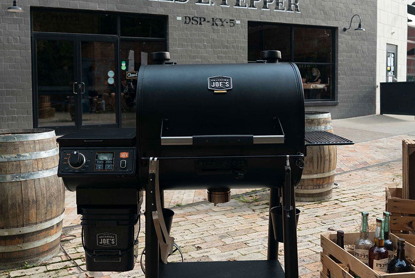 Scenic photograph of the Oklahoma Joe's&reg; Rider DLX Pellet Grill surrounded by rustic barrels and wooden crates.