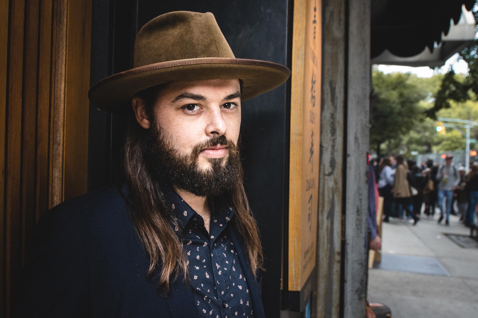 The cover for Caleb Caudle's album titled 'Better Hurry Up' depicting the artist seated in a chair while wearing a hat and sunglasses.